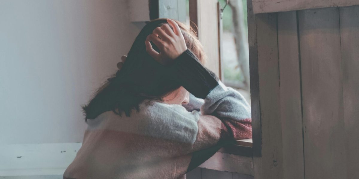 A woman sits indoors gazing thoughtfully out of a window, evoking emotions of loneliness and reflection.
